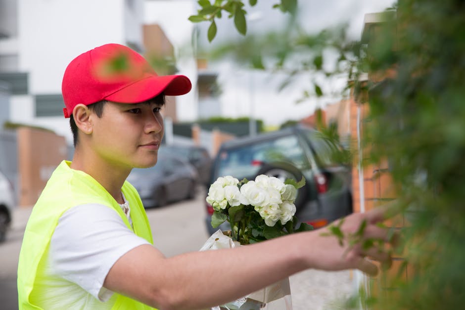 A young male florist wearing a red cap and a yellow high-visibility vest is delivering a floral gift. He is holding a bouquet of fresh white hydrangeas with dark green leaves, wrapped in clear celloph