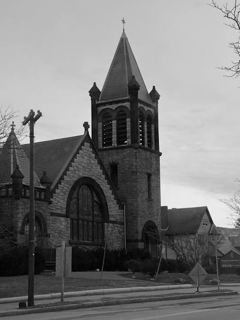 This black-and-white photograph features an old stone church with a tall, pointed steeple topped by a weather vane, situated on a street corner. The church's architecture includes arched windows and a