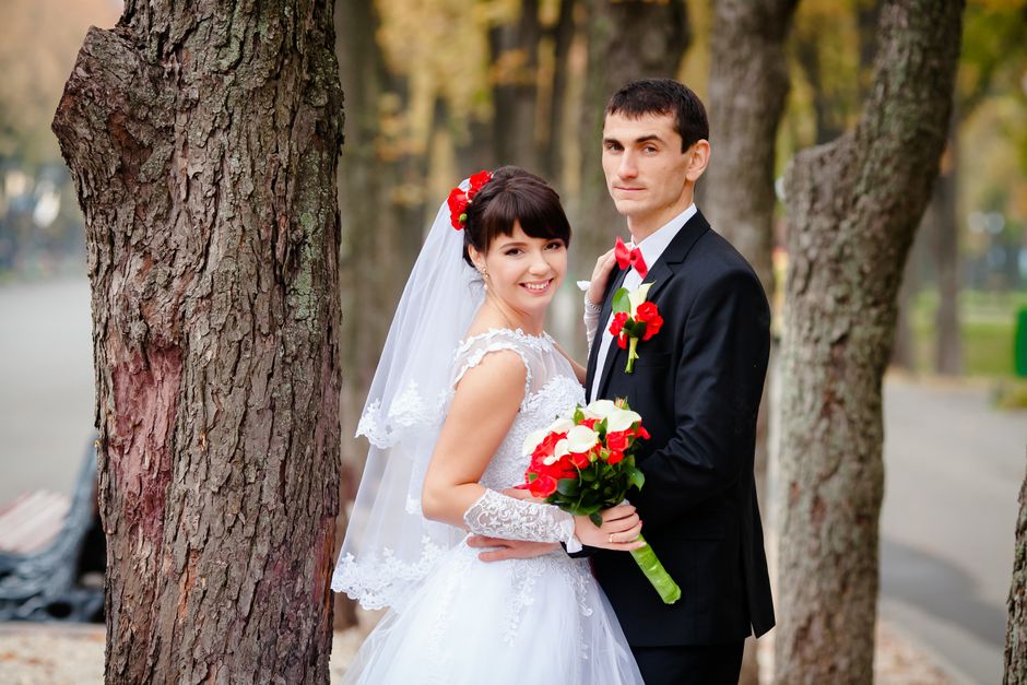 A bride and groom posing outdoors among tall trees with textured bark during a wedding photoshoot. The bride is wearing a white lace wedding dress with delicate cap sleeves, a sheer veil with lace tri