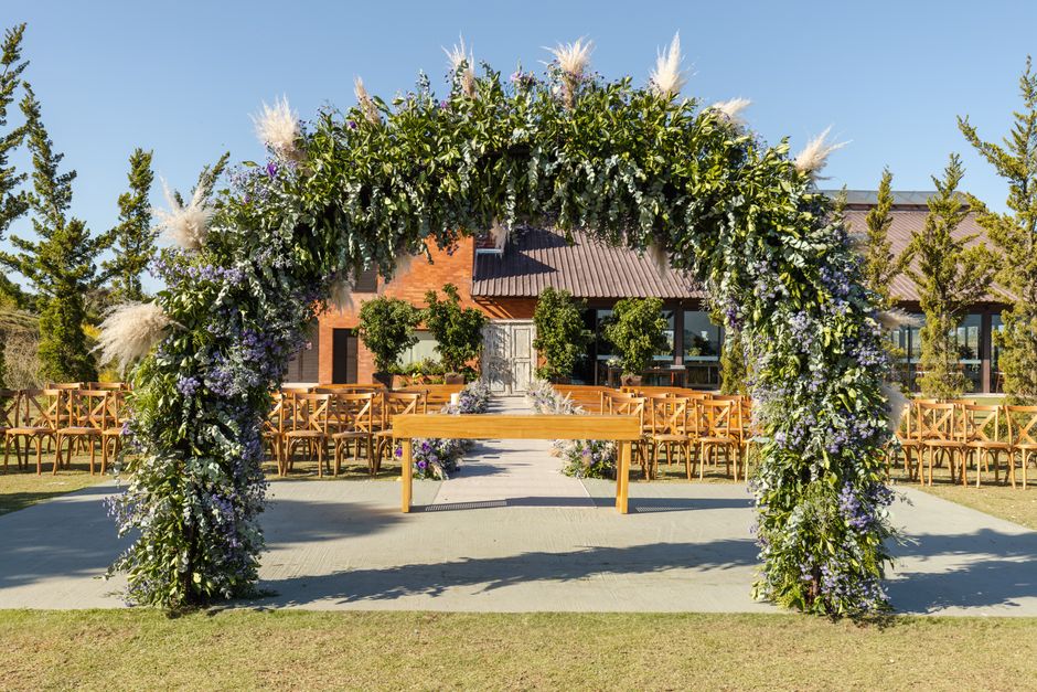 A large floral arch decorated with a lush arrangement of white, purple, and green flowers, including pampas grass, positioned outdoors on a paved area in front of a modern brick building with large wi