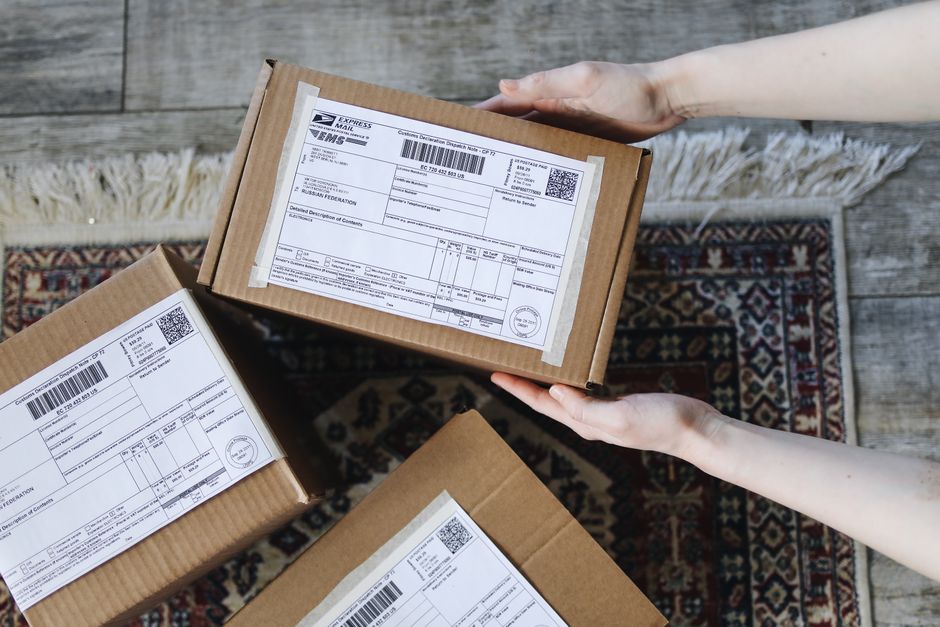 A pair of hands holding a cardboard box with a flower delivery label, shown from above. The box is on a wooden floor with a patterned rug nearby, and other similar parcels with shipping labels are vis