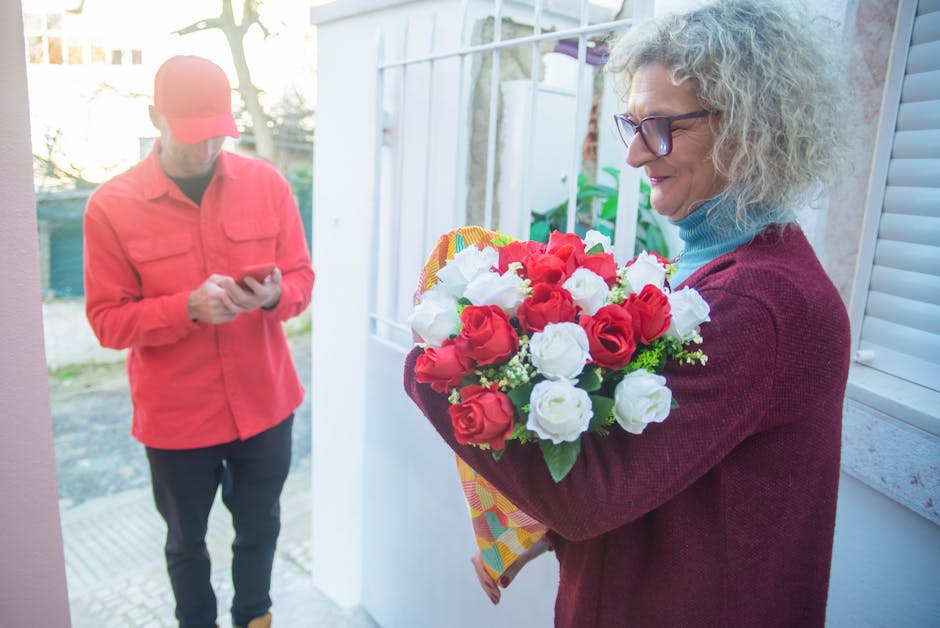 A woman with curly, gray hair and glasses holds a vibrant bouquet of fresh red and white roses wrapped in colorful patterned paper, smiling warmly at the floral gift from Belmont flowers. She is weari