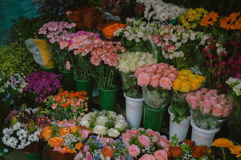 A vibrant display of fresh floral bouquets and potted flowers arranged on a market stand, showcasing a variety of flowers including pink and white roses, orange and yellow roses, white daisies, purple