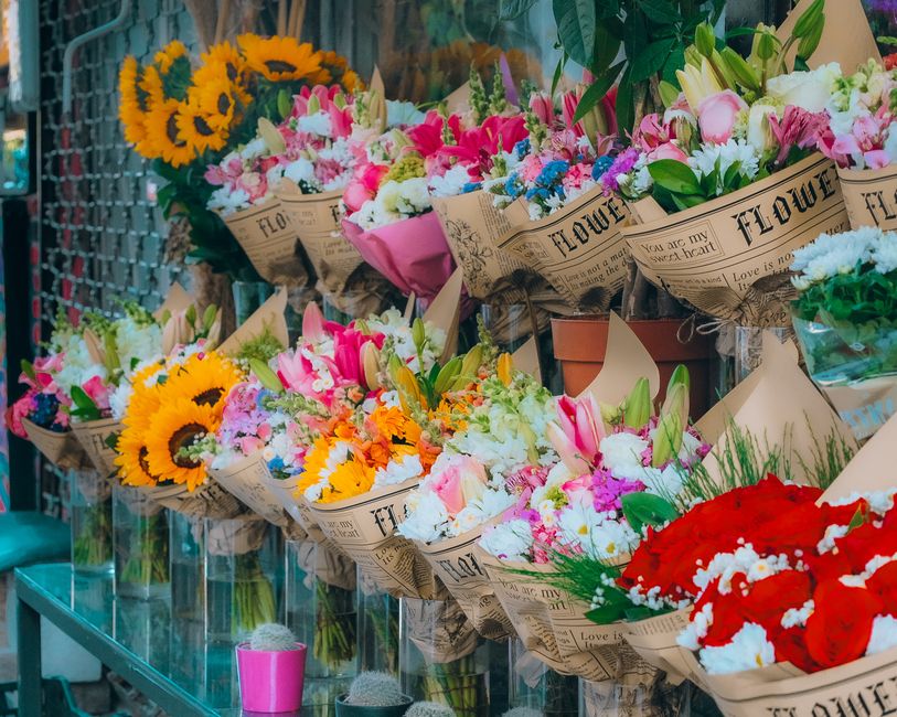 An assortment of fresh floral bouquets arranged on a glass shelf, featuring a variety of vibrant flowers including bright yellow sunflowers, pink and white lilies, blue and purple blooms, and red rose